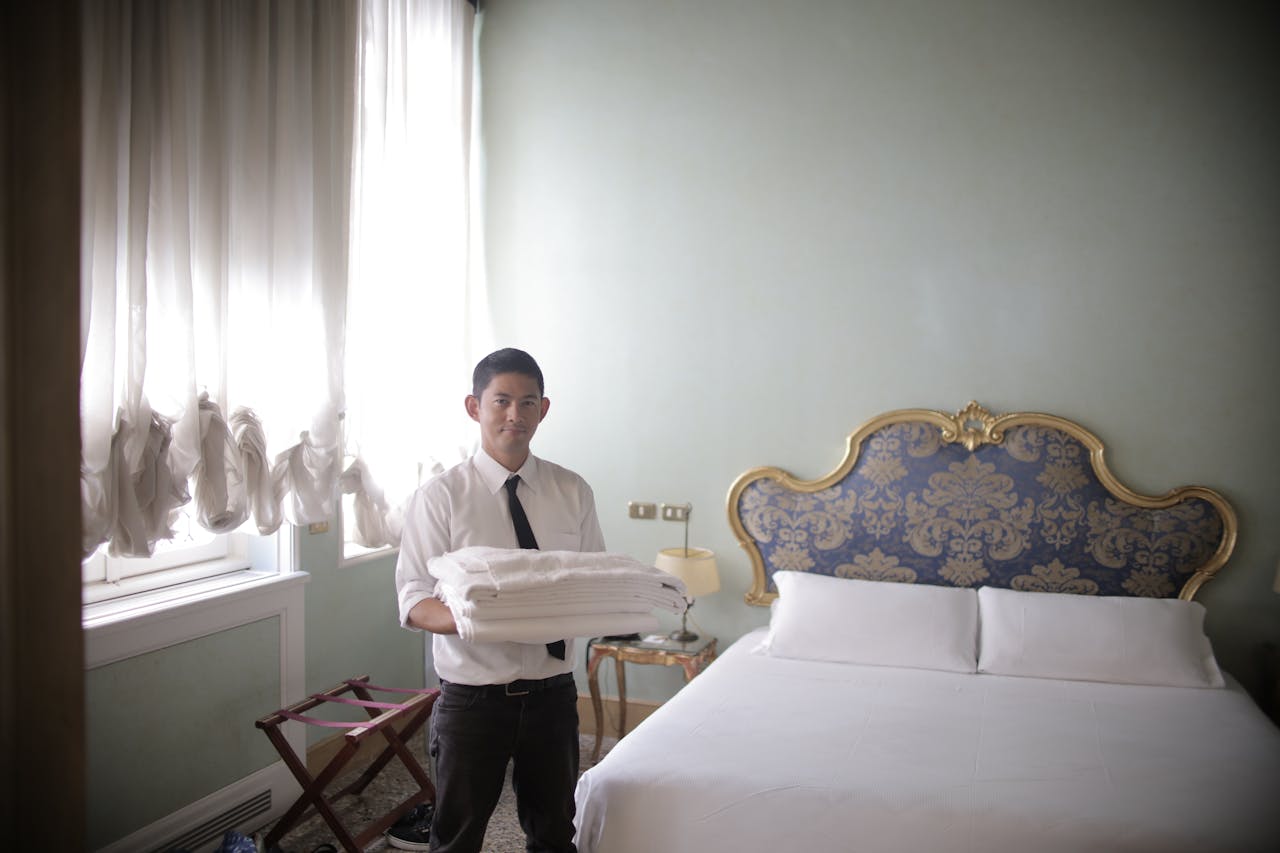 Young ethnic man holding stacked bed linen and looking at camera while preparing comfortable hotel room for guests